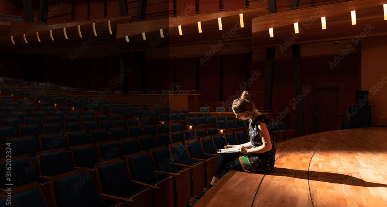 Woman sitting on a stage in a dark theater looking at a clipboard.
