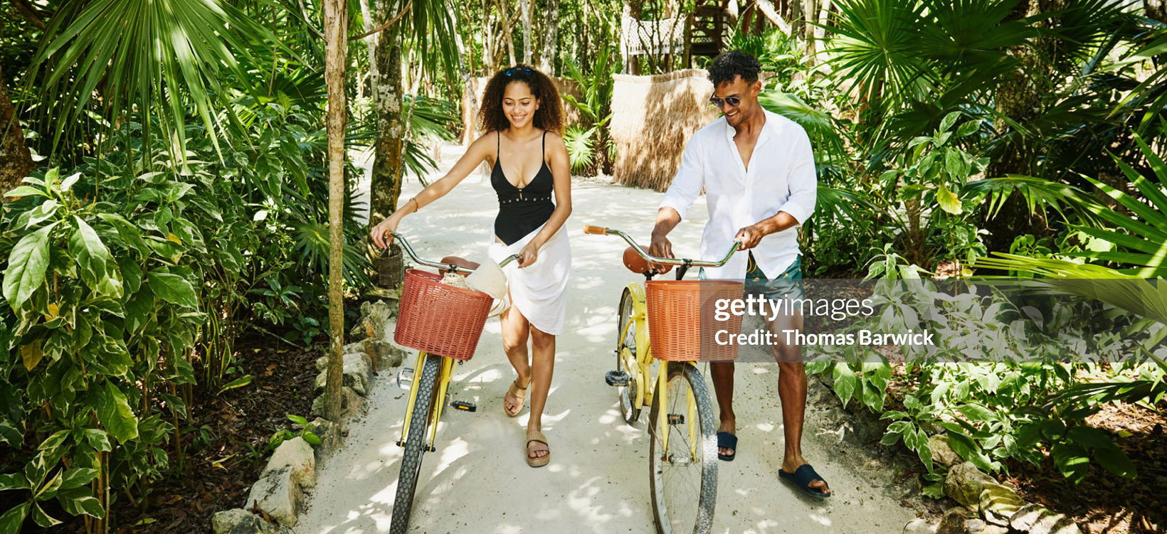 Smiling couple walking bikes on pathway in jungle at tropical resort.